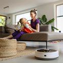 Witte robotstofzuiger met zwarte bovenkant, naast een rieten mand in een woonkamer. Een moeder en kind spelen op de achtergrond.
