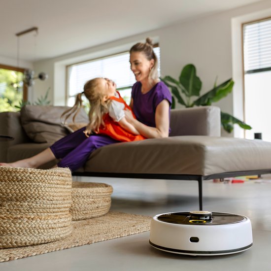 Witte robotstofzuiger met zwarte bovenkant, naast een rieten mand in een woonkamer. Een moeder en kind spelen op de achtergrond.
