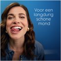 Een jonge vrouw met krullend bruin haar en een blauw shirt likt haar lippen. Haar witte tanden zijn zichtbaar. Tekst: Voor een langdurig schone mond.