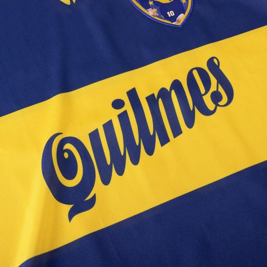 Detail van een blauw voetbalshirt met een opgestikte gele band met de tekst 'Quilmes'.