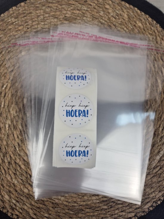 Rol witte stickers met blauwe 'hiep hiep HOERA!' tekst en blauwe stipjes, naast transparante celofaan zakjes met rode plakrand.