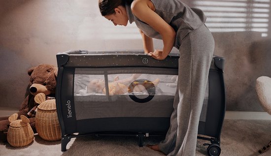 Grijze box met sterrenprint, een baby erin en een vrouw die erbij staat. Grote knuffelbeer en manden op de achtergrond.