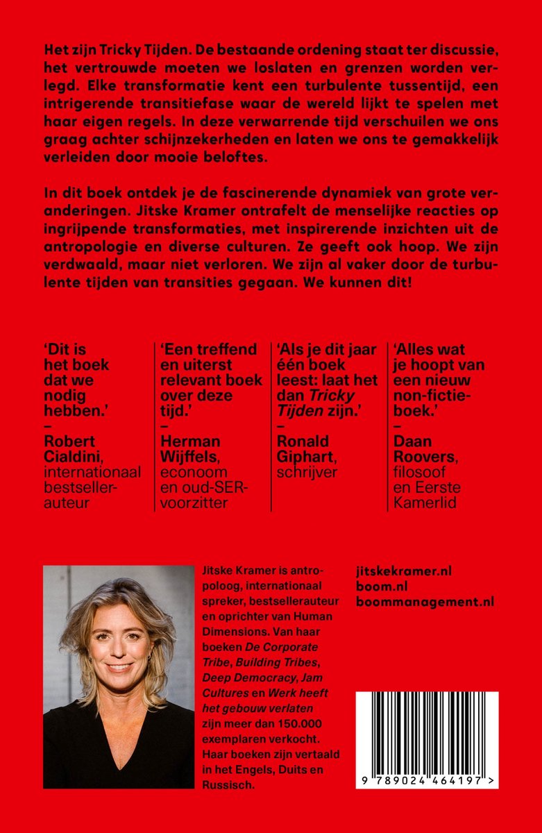 Een vrouw met een open glimlach, met blond haar en bruine ogen, staat op de foto. Ze draagt een zwarte trui. Op de achtergrond een rode poster met tekst.