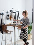 Vrouw met blond paardenstaartje in geblokt schort, staat bij een tafel met ontbijt. Keukendoeken met blokjespatroon hangen aan de muur.