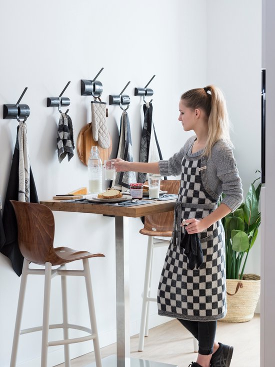 Vrouw met blond paardenstaartje in geblokt schort, staat bij een tafel met ontbijt. Keukendoeken met blokjespatroon hangen aan de muur.