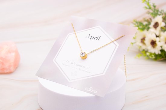 Gouden ketting met zirkonia hanger op een kaart met 'April', naast roze en paarse kristallen.