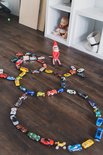 Elf met kerstmuts, rood shirt en gestreepte broek, zittend op een houten vloer omringd door speelgoedauto's.