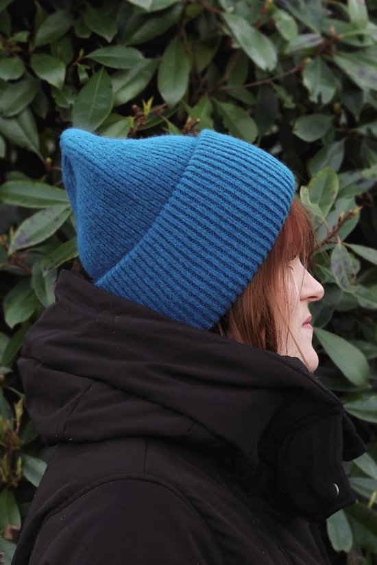 Vrouw van opzij gezien met een blauwe gebreide muts en zwarte jas, met groene bladeren op de achtergrond.