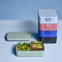 Pile de cinq boîtes à lunch de différentes couleurs : vert, bleu clair, rose, bleu et noir. Une boîte verte est ouverte, avec des sandwichs et de la salade.