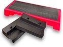 Een rode aerobic step met zwart antislipoppervlak, met twee losse zwarte verhogers ernaast.