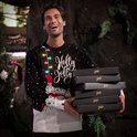 Man met een kersttrui met lichtjes en de tekst "Holly Jolly". Hij draagt de trui met een witte onderlaag en houdt zes zwarte dozen vast.