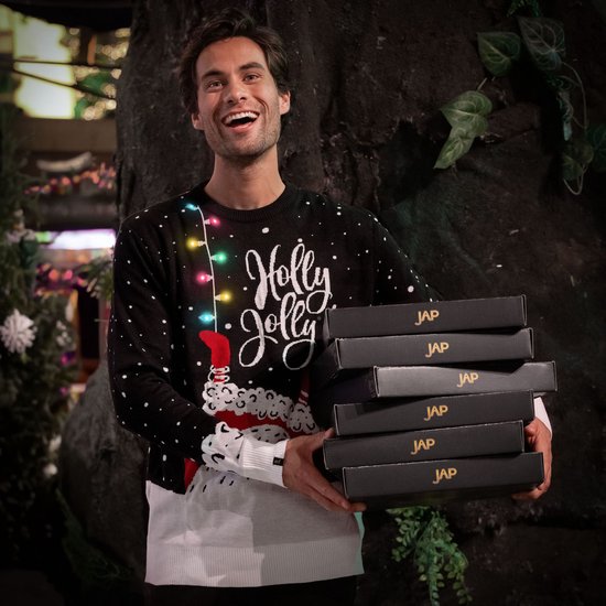 Man met een kersttrui met lichtjes en de tekst "Holly Jolly". Hij draagt de trui met een witte onderlaag en houdt zes zwarte dozen vast.