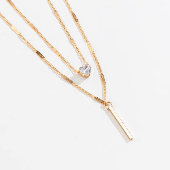 Gouden dubbele ketting met zirkonia hartje en staafje, op witte achtergrond.