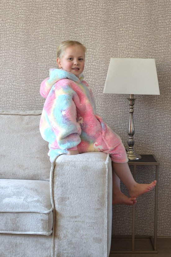 Een jong meisje met blonde staartjes, zittend op de armleuning van een bank, draagt een pluche hoodie deken met pastel regenboogkleuren, sterren en eenhoorns.