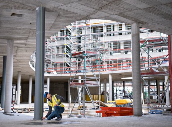 Man in bouwhelm en geel hesje meet een pilaar met Bosch laser afstandmeter. Constructie met steigers op achtergrond.