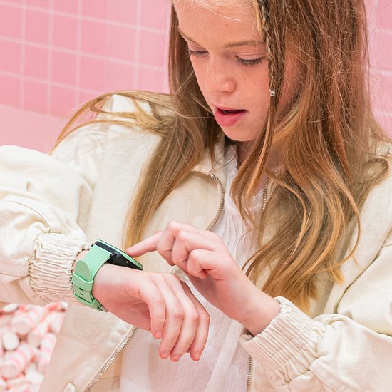 Meisje met blonde vlecht en beige jasje wijst naar een lichtgroene smartwatch om haar pols.