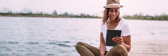 Vrouw met hoed leest op een PocketBook InkPad 4 e-reader op een houten pier aan het water.