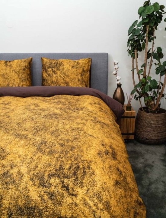 Een grijs hoofdbord met een goudkleurig dekbedovertrek en kussenslopen met een gemarmerd patroon. Een plant staat naast het bed.