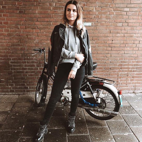 Vrouw in grijze hoodie, zwarte leren jas, zwarte skinny jeans en stoere veterschoenen, staand naast een bromfiets.
