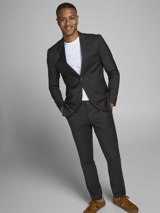 Jonge man met donkere huid draagt een zwarte Jack & Jones blazer met een wit T-shirt en zwarte broek, handen in de zakken.