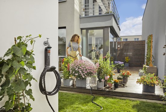 Vrouw met spuitmond op tuinslang, buiten bij bloemen en een opgerolde zwarte tuinslang aan een Gardena houder.