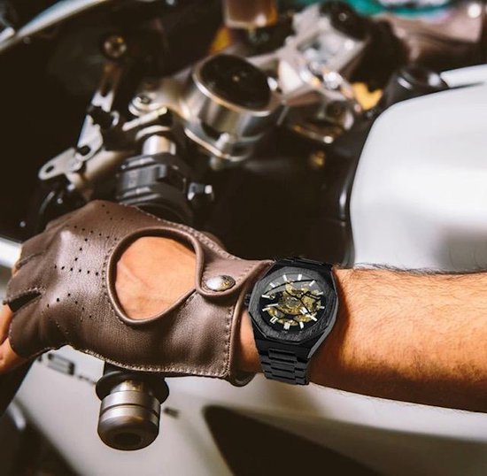 Man met leren handschoenen aan, draagt een zwart automatisch skeleton horloge met gouden tandwielen om de pols, bij een motor.