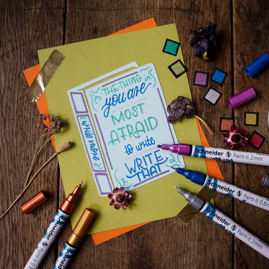 Gekleurde metallic stiften en gedroogde bloemen liggen rond een kaart met de tekst "THE THING you are MOST AFRAID to write write more THAT".
