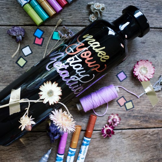 Zwarte fles met tekst "make your holiday metallic" geschreven met metallic stiften, omringd door gedroogde bloemen en fournituren.