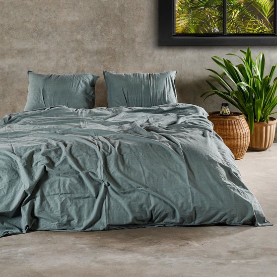 Een tweepersoonsbed met een groen dekbedovertrek en twee kussens, opgemaakt in een slaapkamer met betonnen muur.