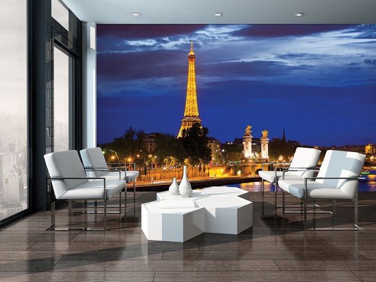 Papier peint représentant la Tour Eiffel illuminée à Paris la nuit, installé dans un salon moderne avec des chaises blanches.