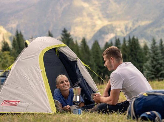 Een stel geniet van koffie bij de ingang van een Coleman Darwin 2 tent in een bergachtige omgeving.