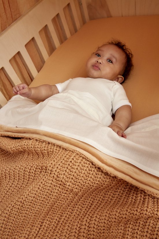 Baby met donkere huidskleur ligt op een wiegje met een oranje laken en een bruin gebreide deken.