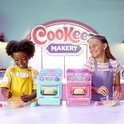 Twee kinderen met keukenschorten lachen terwijl ze naast een turquoise en een roze oven zitten. Een jongen met gele schort mengt iets, een meisje met paarse schort mengt ook.