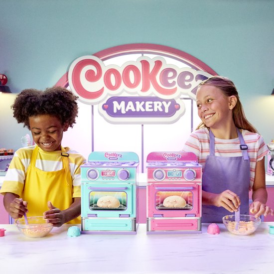 Twee kinderen met keukenschorten lachen terwijl ze naast een turquoise en een roze oven zitten. Een jongen met gele schort mengt iets, een meisje met paarse schort mengt ook.