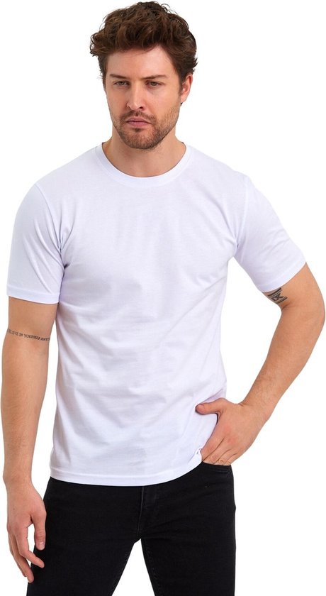 Un homme aux cheveux foncés et à la barbe porte un t-shirt blanc et un pantalon noir. Il a un tatouage sur le bras gauche.