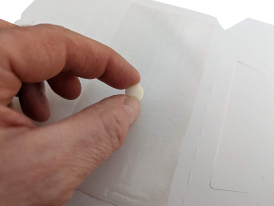 Een hand houdt een kleine, ronde, witte tablet vast boven een open, witte kartonnen doos.