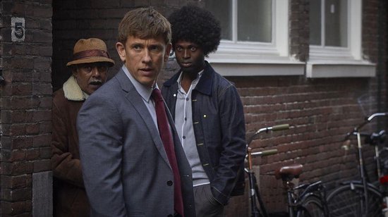 Man in grijs pak met rode das, met een man in spijkerjack en een man met hoed op de achtergrond. Stenen muur.