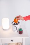 Hand voegt een potje met oranje babyvoeding toe aan de witte flesverwarmer "Thermup 2.0". Op tafel staan ook een potje rode jam en een lamp.