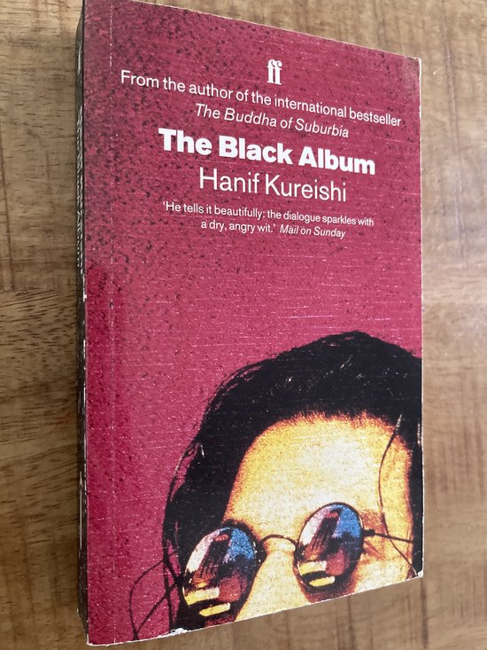 Boek met rode kaft, 'The Black Album' van Hanif Kureishi. Met een close-up van een gezicht met bril.