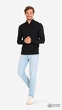 Man staat met zijn handen voor zijn buik in een zwarte trui met rits en lichtblauwe jeans.