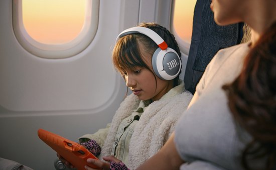 Meisje met gevlochten haar draagt witte en grijze JBL koptelefoon met oranje accenten op een vliegtuig.