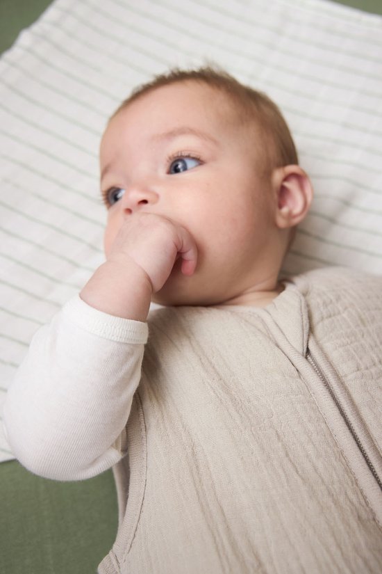 Baby met blauwe ogen, slurpt op duim, draagt een beige slaapzak.