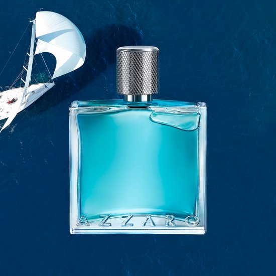 Een Azzaro Chrome Legend parfumfles drijft op de oceaan, met een zeilboot op de achtergrond.