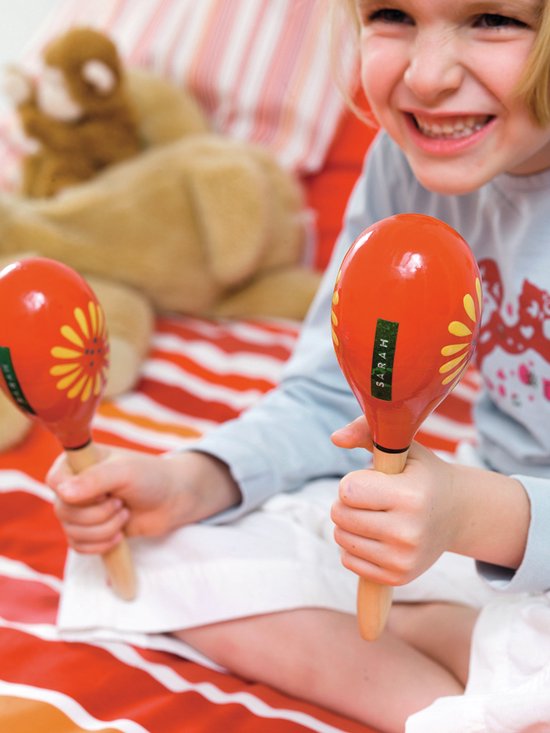 Lachend meisje met blonde haren, houdt twee oranje maracas vast met gele bloemmotieven en "SARAH" erop.