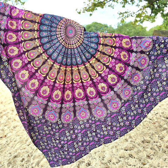 Paars strandkleed met mandala-patroon in roze, geel en cyaan. Hangt aan een boomtak boven het zand.