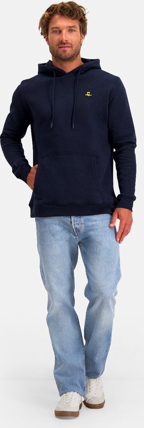 Man met blauwe jeans en donkerblauwe hoodie met gele opdruk op de borst, hand in zak.