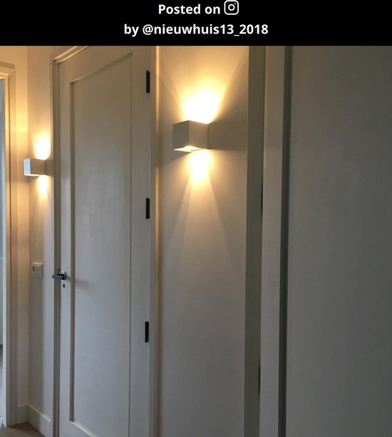 Witte wandlamp op een muur naast een deur, met een brede lichtstraal omhoog en omlaag.