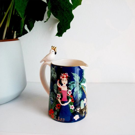 Pitcher met Frida Kahlo print, met bloemen en gebladerte, naast een plant. Witte papegaai met gouden kroon op het handvat.