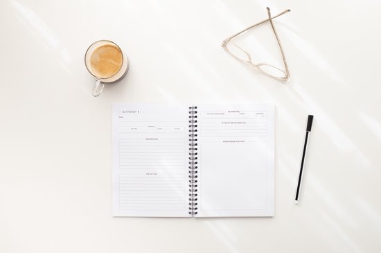 Open notitieboek met ingevulde tekst, kopje koffie, bril en pen op een witte tafel.