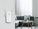 TP-Link RE650 WiFi-repeater in een woonkamer, aangesloten op de muur, naast een grijze bank en salontafel met plant.